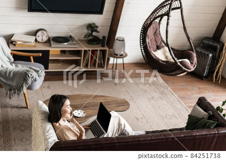 Portrait of young woman sitting at home with laptop. Asian girl using computer pc in her living room, resting on sofa Portrait of young woman sitting at home with laptop. Asian girl using computer pc in her living room, resting on sofa 84251738