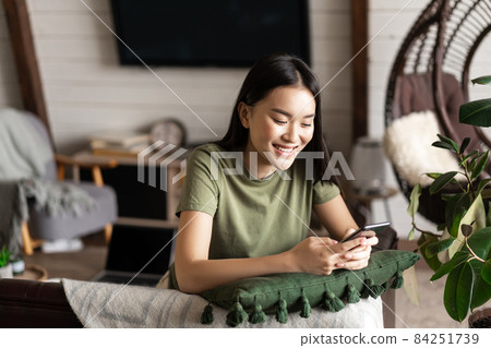 Smiling korean girl chatting on mobile phone, sitting at home in living room with smartphone and laughing happy Smiling korean girl chatting on mobile phone, sitting at home in living room with smartphone and laughing happy 84251739