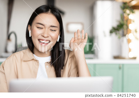 Happy asian woman laughing and smiling, talking on video call using laptop, waving hand and chuckling, wearing earbuds 84251740