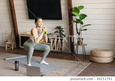 Young asian woman doing workout at home with dumbbels, making squats on floor mat, follow online fitness instructor guide on laptop Young asian woman doing workout at home with dumbbels, making squats on floor mat, follow online fitness instructor guide on laptop 84251741
