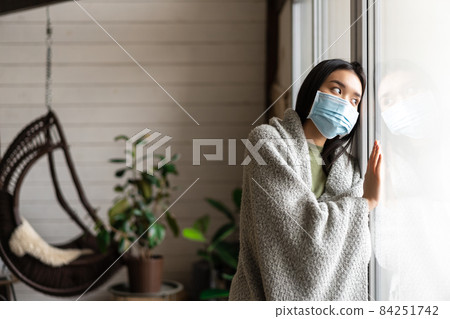 Sad asian girl in medical face mask looking outside window and yearning go for walk, being ill, catching covid 19 or flu 84251742