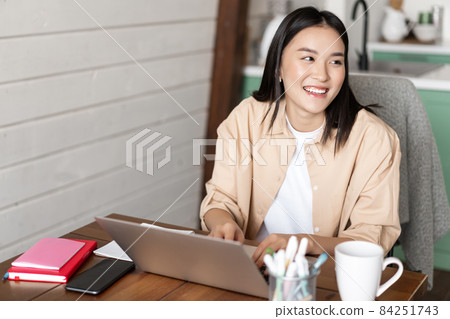 Young asian woman working from home on laptop, studying or doing homework with computer pc, sitting at kitchen table and smiling aside 84251743