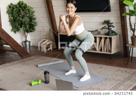Cute asian fitness girl at home doing workout, squats with stretching elastic rope on legs, standing on floor mat, listening to online sport instructor on laptop video 84251745