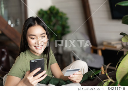 Smiling asian girl sitting at home with mobile phone and credit card, order in online shop, paying for delivery 84251746