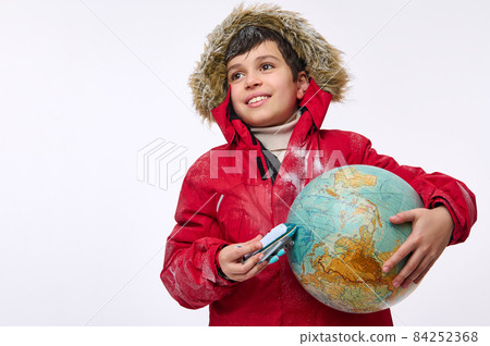 Around the world travel and tourism concept. Adorable elementary aged school boy in warm bright red down jacket with hood holding an airplane toy model and globe, imitating the flight around the Earth Around the world travel and tourism concept. Adorable elementary aged school boy in warm bright red down jacket with hood holding an airplane toy model and globe, imitating the flight around the Earth 84252368