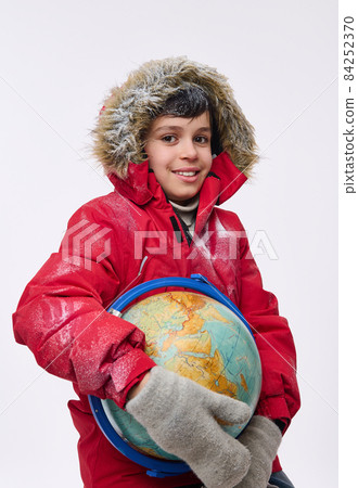 Adorable handsome boy in bright red snow covered down jacket and warm woolen gloves gloves hugging a globe, smiles toothy smile looking at camera. Winter tourism concept with copy ad space Adorable handsome boy in bright red snow covered down jacket and warm woolen gloves gloves hugging a globe, smiles toothy smile looking at camera. Winter tourism concept with copy ad space 84252370