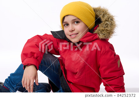 Beautiful child, handsome preadolescent European boy in bright yellow hat and warm red down jacket smiles toothy smile looking at camera sitting on a white background with copy space for advertisement Beautiful child, handsome preadolescent European boy in bright yellow hat and warm red down jacket smiles toothy smile looking at camera sitting on a white background with copy space for advertisement 84252372
