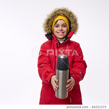 Handsome cheerful school aged boy in bright warm clothes smiles toothy smile looking at camera holding a vacuum flask in front of him, isolated on white background with copy space. Winter trip concept Handsome cheerful school aged boy in bright warm clothes smiles toothy smile looking at camera holding a vacuum flask in front of him, isolated on white background with copy space. Winter trip concept 84252373