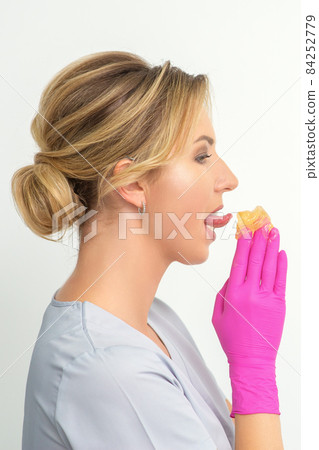 Young beautician licks liquid wax for depilation holding in gloved hands standing on white background. Cosmetologist tastes the sugar paste to taste. 84252779
