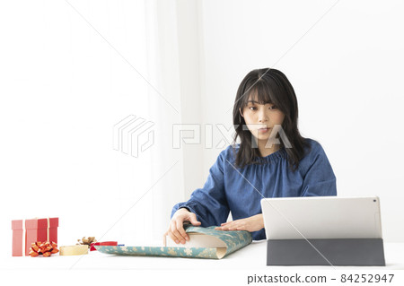 一個女人用包裝紙把禮物包起來，一邊在網上搜索如何把它包在靠近窗戶的桌子上 84252947