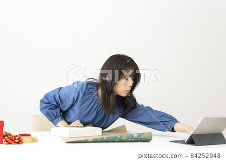 A woman who wraps a gift in wrapping paper while searching the net for how to wrap it on a table near the window 84252948