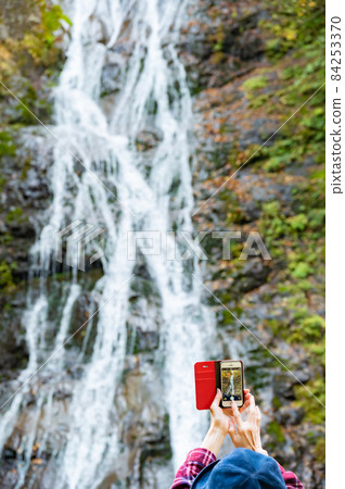 A middle-aged woman taking a picture of the autumn leaves waterfall with a smartphone 84253370