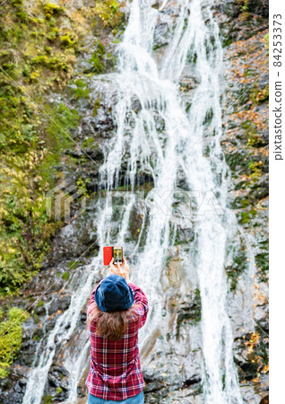 A middle-aged woman taking a picture of the autumn leaves waterfall with a smartphone 84253373