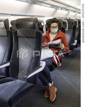 Woman in red duffle coat and protective mask reads magazine in suburban train. Travel by land vehicle. New normal. 84254141