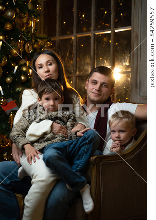 Portrait of a young family. Young parents together with their two sons are sitting on a chair against the background of a Christmas tree Portrait of a young family. Young parents together with their two sons are sitting on a chair against the background of a Christmas tree 84259557