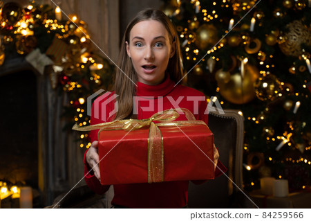 Happy woman in a red sweater opens a magical Christmas gift at home on the background of a Christmas tree 84259566
