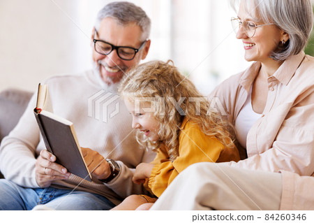 Cute little girl granddaughter smiling when reading book with senior grandparents 84260346