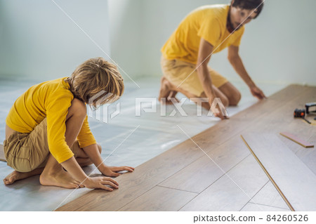 Father and son installing new wooden laminate flooring on a warm film floor. Infrared floor heating system under laminate floor Father and son installing new wooden laminate flooring on a warm film floor. Infrared floor heating system under laminate floor 84260526