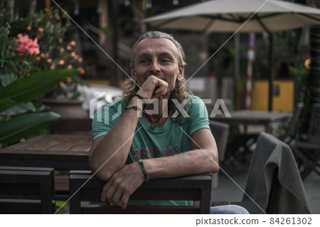 Portrait of mature handsome man sitting on the summer veranda of the restaurant, holding his hand to his chin. Long grey hair. High quality photo 84261302
