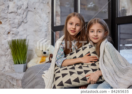 The image of two cute little sisters sitting on windowsill, covered by blanket, hugging each other The image of two cute little sisters sitting on windowsill, covered by blanket, hugging each other 84265635
