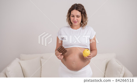 A pregnant woman is holding an apple and a bar of chocolate. The ninth month of pregnancy. 84266518