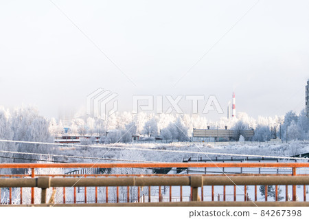 The cityscape is a frosty morning after a snowfall. Boiler room pipes, bridge, buildings, snow-covered trees. Russia, winter. 84267398