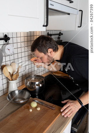 A man cooks broth in the kitchen and tastes it. Household chores. Front view. A man cooks broth in the kitchen and tastes it. Household chores. Front view. 84267399