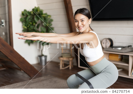 Cute asian fitness girl at home doing workout, squats with stretching elastic rope on her legs, exercising for fit and healthy body Cute asian fitness girl at home doing workout, squats with stretching elastic rope on her legs, exercising for fit and healthy body 84270754