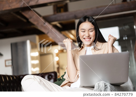 Triumphing asian girl winning online, celebrating victory while watching video on laptop at home 84270779