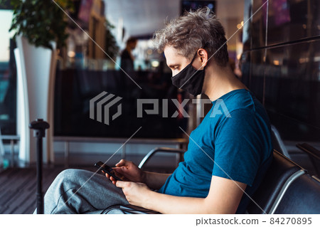 Man wearing face mask and using his phone while waiting for flight at airport terminal. New normal, digital nomad and freelance lifestyle. Traveling during pandemic and personal protection. 84270895