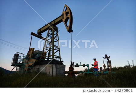 Mechanic in work vest and construction helmet standing near pipeline, to the left of pump jack and recording indicators of oil rig work on the background of clear blue sky. Mechanic in work vest and construction helmet standing near pipeline, to the left of pump jack and recording indicators of oil rig work on the background of clear blue sky. 84270901