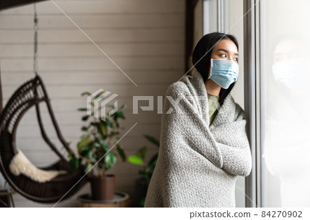 Asian girl getting ill, wearing face medical mask, staying on covid-19 quarantine and looking outside, yearning to go on walk Asian girl getting ill, wearing face medical mask, staying on covid-19 quarantine and looking outside, yearning to go on walk 84270902