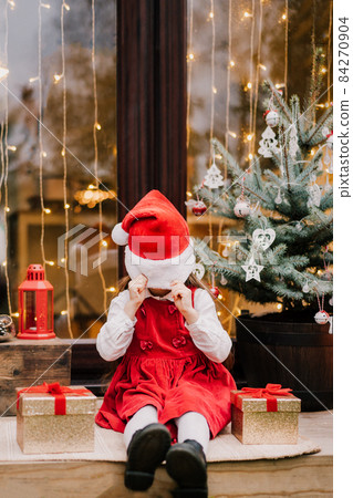 Bored cute little girl kid in red dress pulled Santa Claus hat on her face while sitting with gift boxes on the house porch with decorated Christmas tree. Christmas holiday season. Vertical card. Bored cute little girl kid in red dress pulled Santa Claus hat on her face while sitting with gift boxes on the house porch with decorated Christmas tree. Christmas holiday season. Vertical card. 84270904