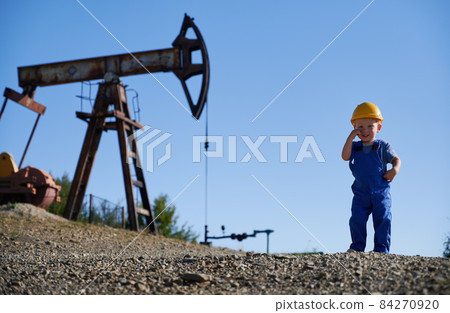 Adorable male child looking at camera and smiling while standing in oil field with petroleum pump jack and blue sky on background. Cute little boy wearing construction helmet and work overalls. 84270920