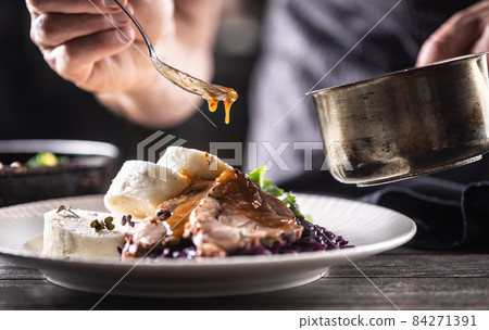 Detail of chef's hand pouring sauce over pork slice with dumplings and red cabbage 84271391