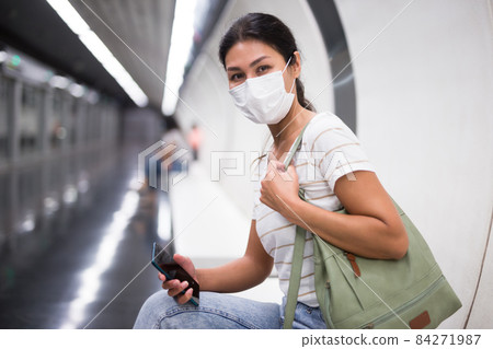 Woman in face mask with smartphone sitting on bench in subway station Woman in face mask with smartphone sitting on bench in subway station 84271987