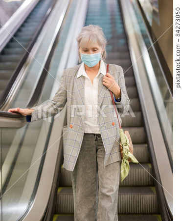 Senior woman in mask on escalator Senior woman in mask on escalator 84273050