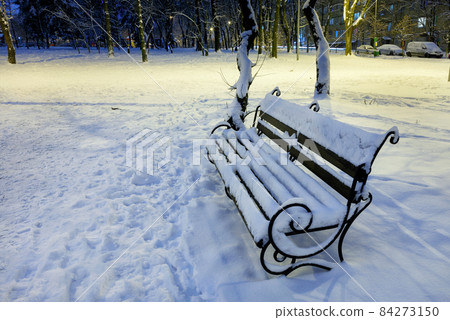 Empty wooden bench covered with snow in a city park in the evening. Empty wooden bench covered with snow in a city park in the evening. 84273150