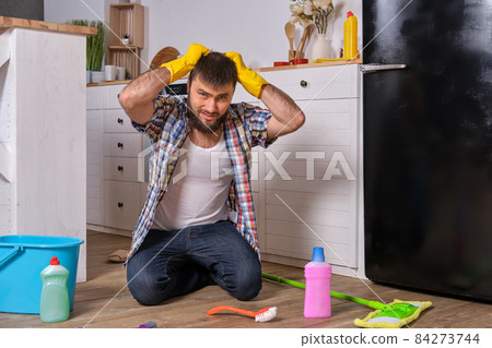Caucasian young bearded man sits on the floor of his kitchen and tries to clean it using all of his detergents, rags, washcloths and brushes 84273744