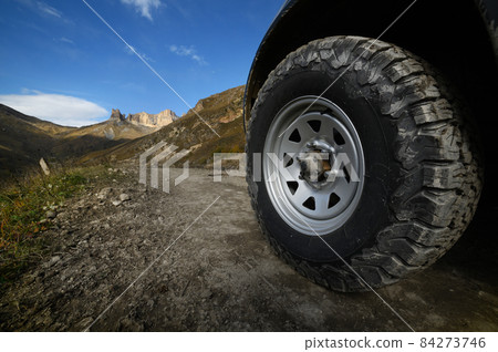 Close-up A large off-road all-terrain car wheel stands on a rough road in the mountains against the backdrop of rocks. Off-road tours and off-road travel concept 84273746
