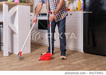 Young handsome bearded man in a kitchen, wearing a plaid shirt, sweeping the floor with a shovel and broom. 84273801