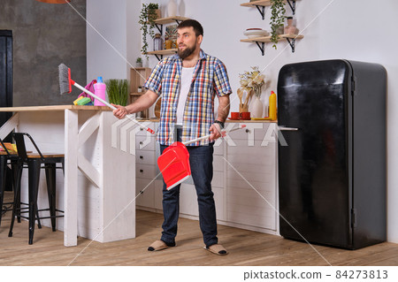 Young handsome bearded man in the kitchen, wearing checkered shirt, tries to deal with a dustpan and a broom. Young handsome bearded man in the kitchen, wearing checkered shirt, tries to deal with a dustpan and a broom. 84273813