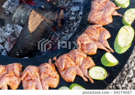 Top above view of many tasty raw marinated quails and vegetables and mushrooms grilled cooked at round steel iron firepit hearth table surface brazier with burning firewoods. Barbecue yard home 84274087