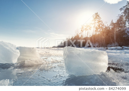 Scenic view of clear ice shard piece on coast of frozen lake landscape against warm sun shining rays through forest tree and clear blue sky on background. Winter weather nature abstract scene view Scenic view of clear ice shard piece on coast of frozen lake landscape against warm sun shining rays through forest tree and clear blue sky on background. Winter weather nature abstract scene view 84274099