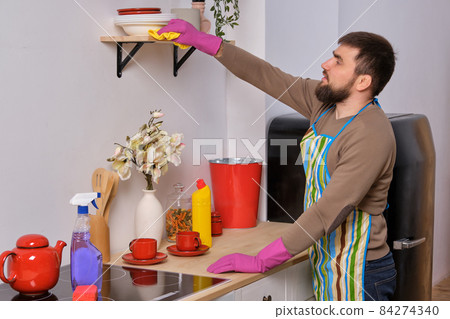 Young handsome bearded man in the kitchen, wearing apron and pink gloves cleans the shelf above the cooking surface using detergents Young handsome bearded man in the kitchen, wearing apron and pink gloves cleans the shelf above the cooking surface using detergents 84274340