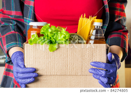A young woman volunteer is holding a donation box with foodstuffs. Delivery of necessary food during an epidemic. A young woman volunteer is holding a donation box with foodstuffs. Delivery of necessary food during an epidemic. 84274805