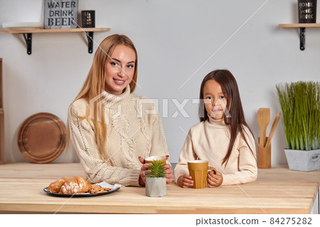 Shot of cheerful mother and daughter sit together at kitchen table, drink hot tea in morning, have pleasant friendly talk between each other. Shot of cheerful mother and daughter sit together at kitchen table, drink hot tea in morning, have pleasant friendly talk between each other. 84275282