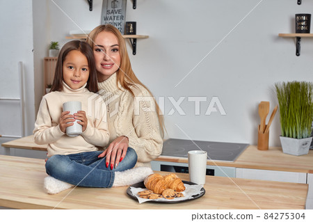 Shot of cheerful mother and daughter sit together at kitchen table, drink hot tea in morning, have pleasant friendly talk between each other. Shot of cheerful mother and daughter sit together at kitchen table, drink hot tea in morning, have pleasant friendly talk between each other. 84275304