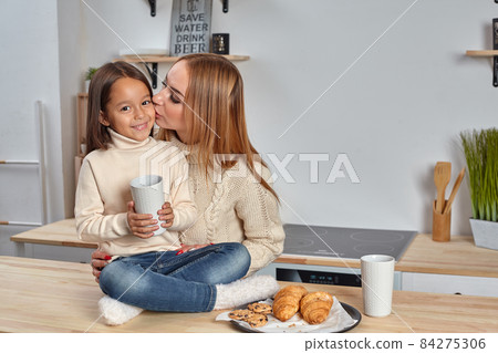 Shot of cheerful mother and daughter sit together at kitchen table, drink hot tea in morning, have pleasant friendly talk between each other. 84275306