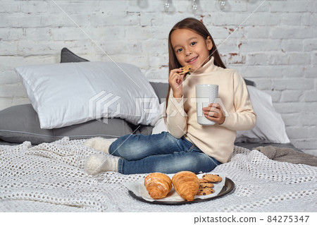 Pretty little girl 6 years old in a white sweater and jeans. Child in the room with a bed, eating croissant and drinking tea or cocoa. Pretty little girl 6 years old in a white sweater and jeans. Child in the room with a bed, eating croissant and drinking tea or cocoa. 84275347